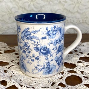 Op de afbeelding: Witte keramische mok met een blauw bloemenpatroon en een blauw interieur. De mok heeft een handvat en is versierd met een gedetailleerd blauw rozenontwerp. De mok staat op een wit kanten kleedje.