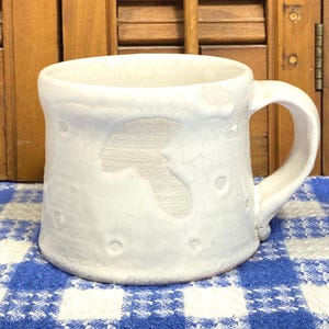 May include: A white ceramic mug featuring a subtle, embossed mushroom silhouette on the side. The surface has a distinct crackle glaze finish and small, circular indentations scattered around the exterior, complete with a sturdy, curved handle.