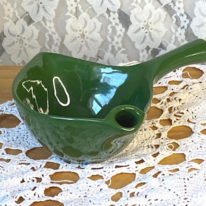 May include: A glossy, emerald green ceramic gravy boat with a handle and a spout. The boat has a rounded shape with a slightly flared rim. The handle is curved and extends from the side of the boat. The spout is small and round.
