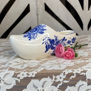 May include: A white ceramic Dutch clog with blue floral designs. The decorative shoe is accented with pink carnations. The clog is displayed on a white lace tablecloth, with a white and black background.
