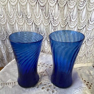 Pu&ograve; includere: Due bicchieri di vetro blu cobalto con un design a spirale. I bicchieri hanno una forma curva e poggiano su una tovaglia di pizzo bianco. Lo sfondo presenta una tenda di pizzo bianco.