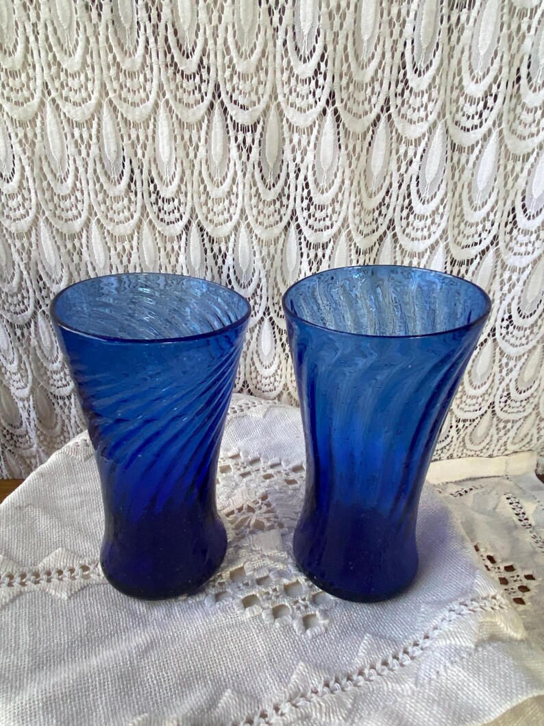 Pu&ograve; includere: Due bicchieri di vetro blu cobalto con un design a spirale. I bicchieri hanno una forma curva e poggiano su una tovaglia di pizzo bianco. Lo sfondo presenta una tenda di pizzo bianco.