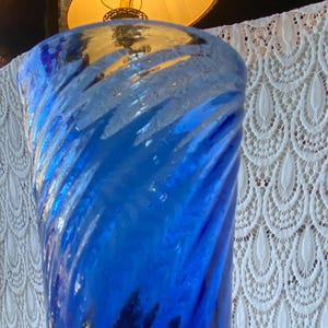 Pu&ograve; includere: Un vaso di vetro blu vibrante con un design a spirale. Il vaso ha una superficie strutturata e una finitura lucida. Lo sfondo include una lampada con un paralume giallo e una tenda di pizzo bianco.