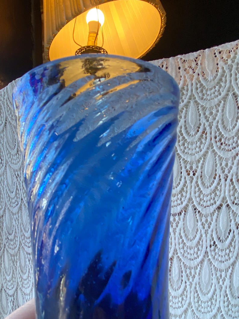 Pu&ograve; includere: Un vaso di vetro blu vibrante con un design a spirale. Il vaso ha una superficie strutturata e una finitura lucida. Lo sfondo include una lampada con un paralume giallo e una tenda di pizzo bianco.