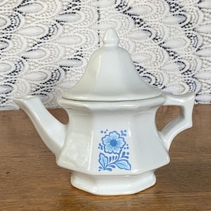 Puede incluir: Una tetera de cerámica blanca con tapa, boquilla y asa. La tetera presenta un diseño floral azul en la parte delantera. La tetera está sobre una superficie de madera.