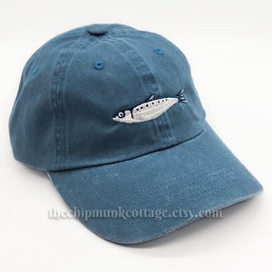 May include: A blue baseball cap with an embroidered white fish design. The cap has a curved brim and a button on top. The text "thechipmunkcottage.etsy.com" is printed on the brim.