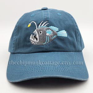 May include: A blue baseball cap featuring an embroidered anglerfish design in gray, white, and teal. The cap has a curved brim and a fabric strap closure. The text "thechipmunkcottage.etsy.com" is printed on the brim.