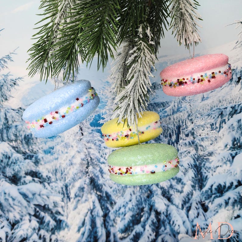 May include: Four colourful macaron ornaments in blue, yellow, pink, and green hang from a frosted evergreen branch. Each ornament has a white filling and is decorated with colourful sprinkles. The background features a snowy winter scene.