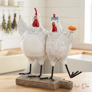 May include: A decorative figurine featuring two white chickens standing on a wooden cutting board. The left chicken wears a white apron, while the right chicken has a blue bow on its head and holds a small orange object. Both have red combs.