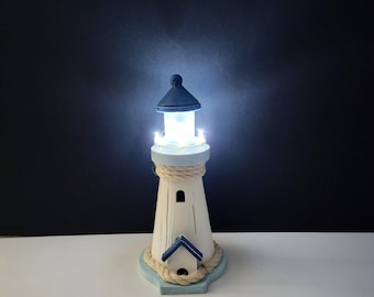 May include: A decorative white wooden lighthouse with a blue roof and base. The lighthouse has a rope detail around the middle and a small house attached. The top of the lighthouse is lit with small white lights.