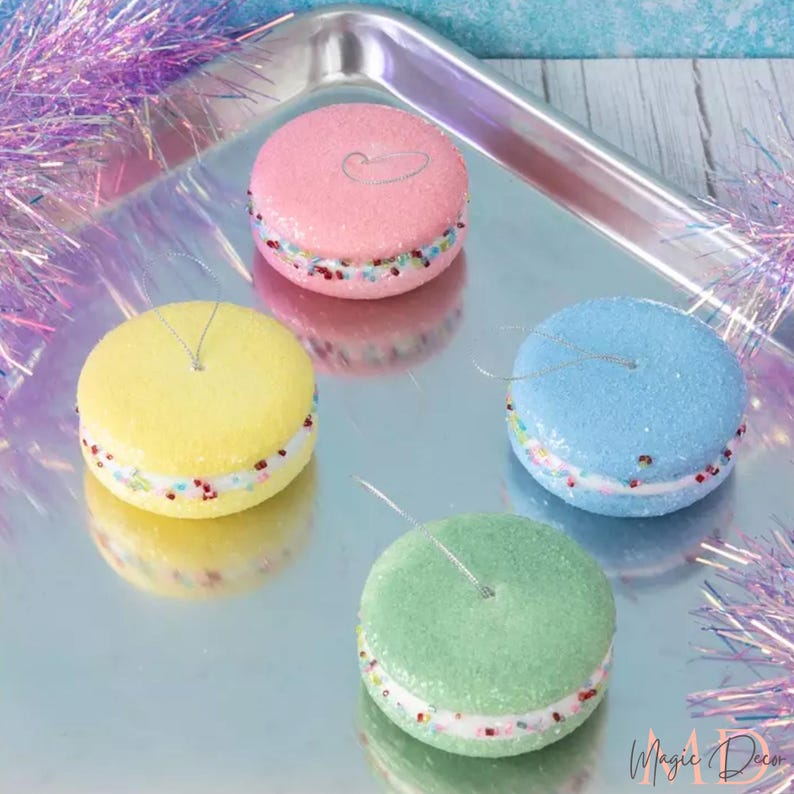 May include: Four colourful macaron ornaments in pink, yellow, blue, and green, each with a sparkly, textured surface and a white filling with colourful sprinkles. Silver string loops for hanging. The ornaments are on a silver tray.