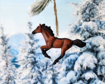 May include: A brown horse ornament hanging from a tree branch. The ornament is made of plastic and has a realistic look. The horse is in a running pose with its head turned to the side. The background is a snowy forest.