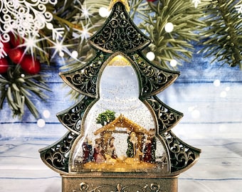 Puede incluir: Un globo de nieve de árbol de Navidad dorado y verde con una escena de la natividad en su interior. El globo de nieve está iluminado con una luz blanca cálida y tiene un efecto de brillo giratorio.