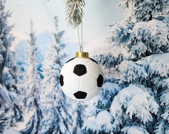 May include: A white and black football bauble with a gold top hanging from a snowy pine tree branch.
