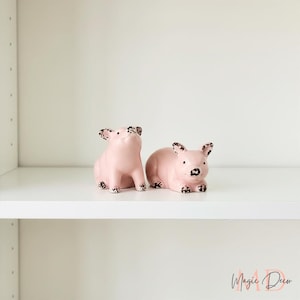 May include: Two pink ceramic pig figurines with black spotted details on their ears, snouts, and feet. One pig is standing, the other is lying down. The figurines are displayed on a white shelf.