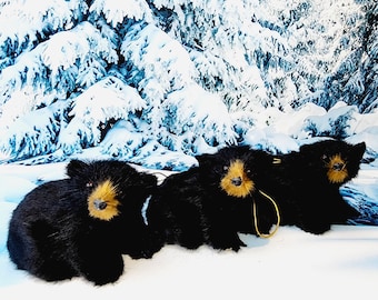 May include: Three black bear ornaments with golden-brown snouts and fur, set against a snowy backdrop of snow-covered trees. The bears are positioned in a row, creating a wintery scene.