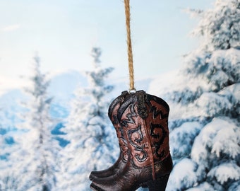 Puede incluir: Un adorno navideño de bota vaquera marrón y negra con un diseño decorativo. El adorno está colgado de una cuerda de yute marrón.