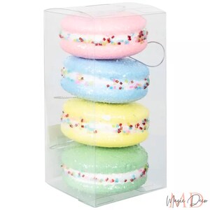 May include: Four decorative macarons in a clear box. The macarons are pastel shades of pink, blue, yellow, and green, with white filling and colourful sprinkles. Each has a silver string for hanging.