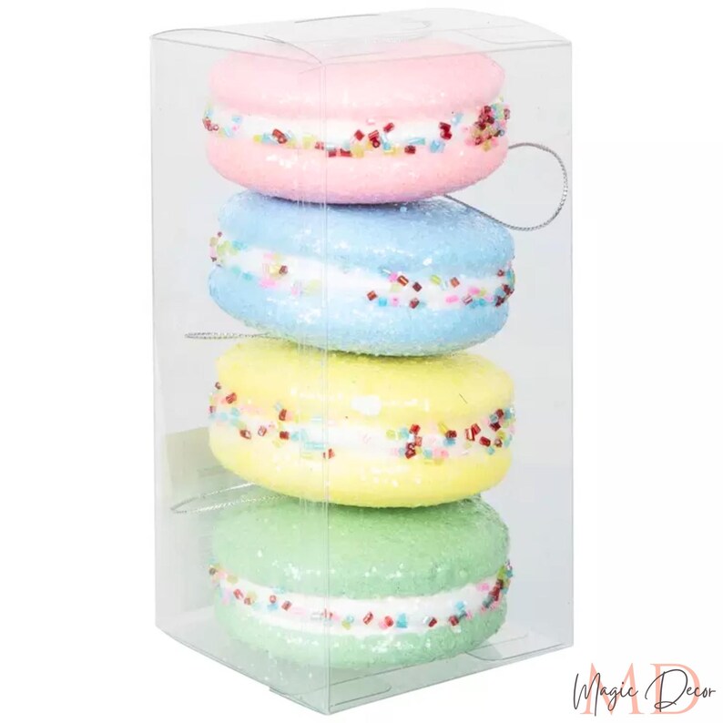 May include: Four decorative macarons in a clear box. The macarons are pastel shades of pink, blue, yellow, and green, with white filling and colourful sprinkles. Each has a silver string for hanging.