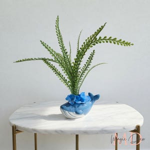 May include: A blue ceramic whale planter with a green plant and blue flower accents. The whale-shaped planter has a white belly and is sitting on a white marble table with gold legs. Decorative home decor.