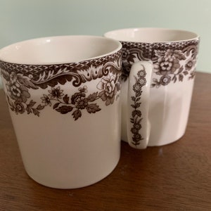 May include: Two white ceramic mugs with brown floral designs. The mugs have a classic shape and a simple design.