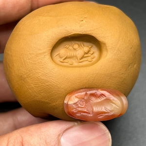 May include: A close-up of a red carnelian gemstone carved with a detailed image of a bull. The gemstone is set in a clay mold, showing the impression of the carving.