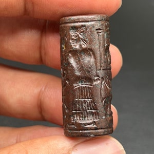 May include: A brown cylindrical seal with intricate carvings depicting a figure with a headdress and other symbols. The seal is made of a hard stone and has a smooth, polished surface.