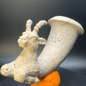 May include: A white ceramic sculpture of a goat with horns, standing on a curved base. The sculpture is resting on a bright orange surface.