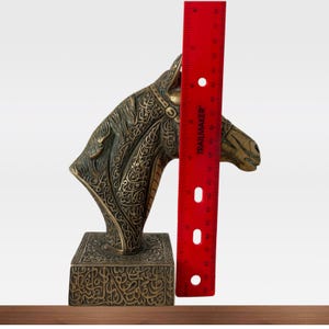 May include: A bronze-colored horse head sculpture with intricate engraved patterns. The sculpture is mounted on a rectangular base with similar engraved designs. A red ruler is placed next to the sculpture, measuring approximately 10 inches in height.