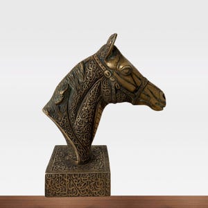 May include: A bronze horse head sculpture with intricate engraved patterns. The sculpture features a detailed bridle and is mounted on a rectangular base, also adorned with engraved designs. The horse head is a rich, antique gold color.