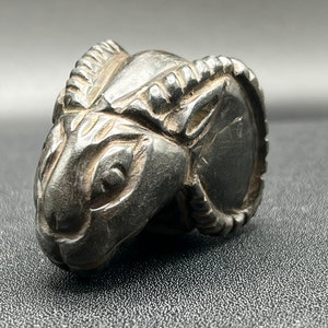 May include: A small, black, carved ram's head sculpture. The ram's head is detailed with horns and facial features.
