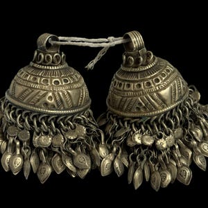 May include: Two silver-toned bell-shaped ornaments with intricate etched patterns. Each bell has a series of dangling leaf-shaped charms. The bells are connected by a white string against a black background.