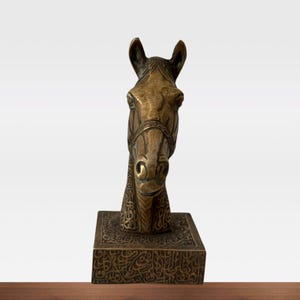 May include: A bronze horse head sculpture with intricate detailing. The horse head is mounted on a rectangular base, also adorned with detailed patterns. The sculpture is a warm, metallic color and appears to be an antique or vintage piece.