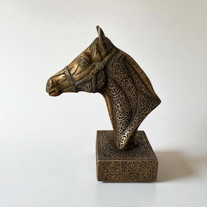 May include: A bronze horse head sculpture with intricate engraved patterns and a bridle. The sculpture is mounted on a square base, also adorned with detailed carvings. The artwork is a decorative piece.