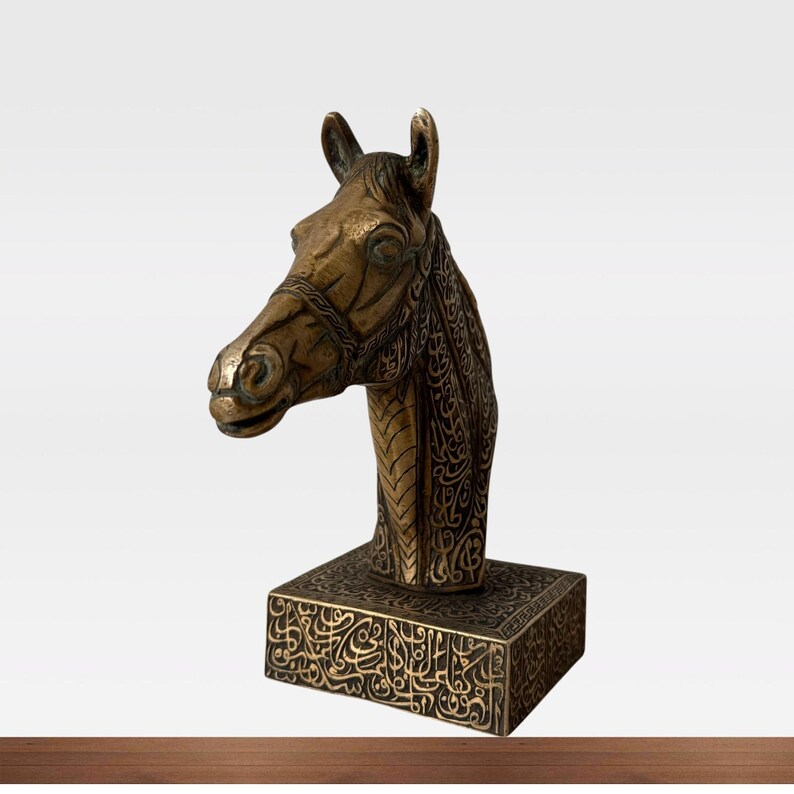 May include: A bronze-colored horse head sculpture with intricate detailing. The horse head is mounted on a square base with ornate patterns. The sculpture has a vintage or antique aesthetic.