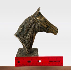 May include: A bronze-colored horse head sculpture with intricate engraved patterns. The sculpture is mounted on a square base and measures approximately 10 inches in length. The horse head is detailed with a bridle and decorative elements.