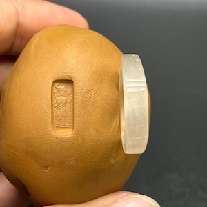 May include: A white jade ring with a carved design. The ring is shown on a brown clay surface.