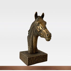 May include: A bronze horse head sculpture with intricate detailing. The sculpture features a horse head with a bridle, mounted on a square base. The base and the horse's neck are adorned with ornate patterns. The sculpture is a decorative item.