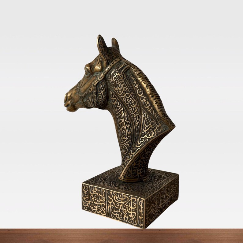 May include: A bronze-colored horse head sculpture with intricate engraved patterns. The sculpture is mounted on a square base, also adorned with detailed carvings. The horse head is depicted in profile, showcasing its elegant features and the ornate bridle.