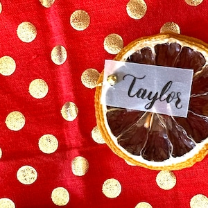 May include: A dried orange slice place card with a white card that says "Taylor" on a red background with gold polka dots.