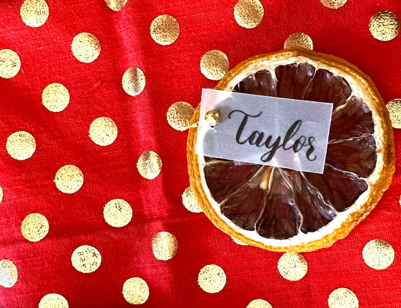 May include: A dried orange slice place card with a white card that says "Taylor" on a red background with gold polka dots.