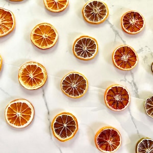 May include: A collection of dried orange and lime slices arranged on a white marble surface. The citrus slices are a variety of colors, including yellow, orange, and brown.