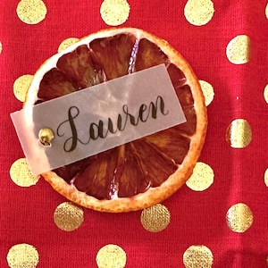 May include: A dried orange slice with a translucent paper place card that says "Lauren" on a red background with gold polka dots.