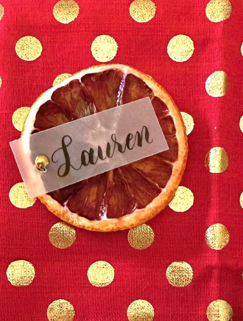 May include: A dried orange slice with a translucent paper place card that says "Lauren" on a red background with gold polka dots.