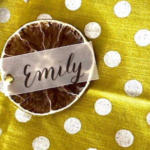 May include: A dried lime slice with a white paper tag that says "Emily" on a yellow fabric background with white polka dots.