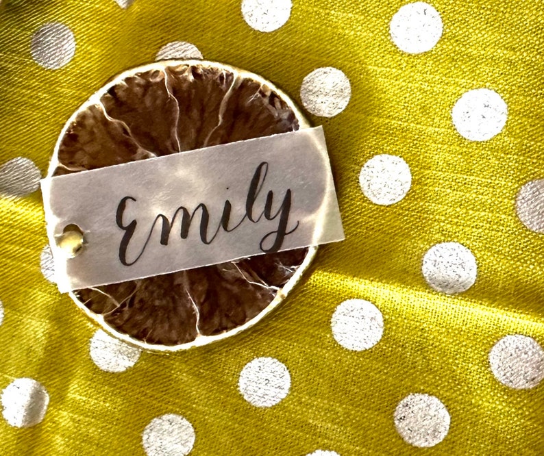 May include: A dried lime slice with a white paper tag that says "Emily" on a yellow fabric background with white polka dots.