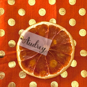 May include: Dried orange slice place card with the name Audrey written on a white tag. The place card is on a orange tablecloth with gold polka dots.