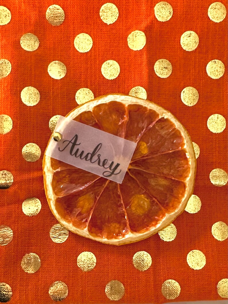 May include: Dried orange slice place card with the name Audrey written on a white tag. The place card is on a orange tablecloth with gold polka dots.
