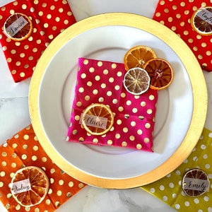 May include: Five place settings with gold rimmed plates and napkins with gold polka dots. Each place setting has a dried orange slice with a name tag on top. The names are: Lauren, Taylor, Claire, Audrey, and Emily.