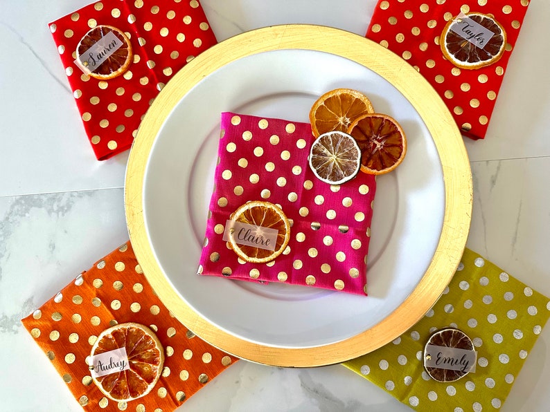 May include: Five place settings with gold rimmed plates and napkins with gold polka dots. Each place setting has a dried orange slice with a name tag on top. The names are: Lauren, Taylor, Claire, Audrey, and Emily.